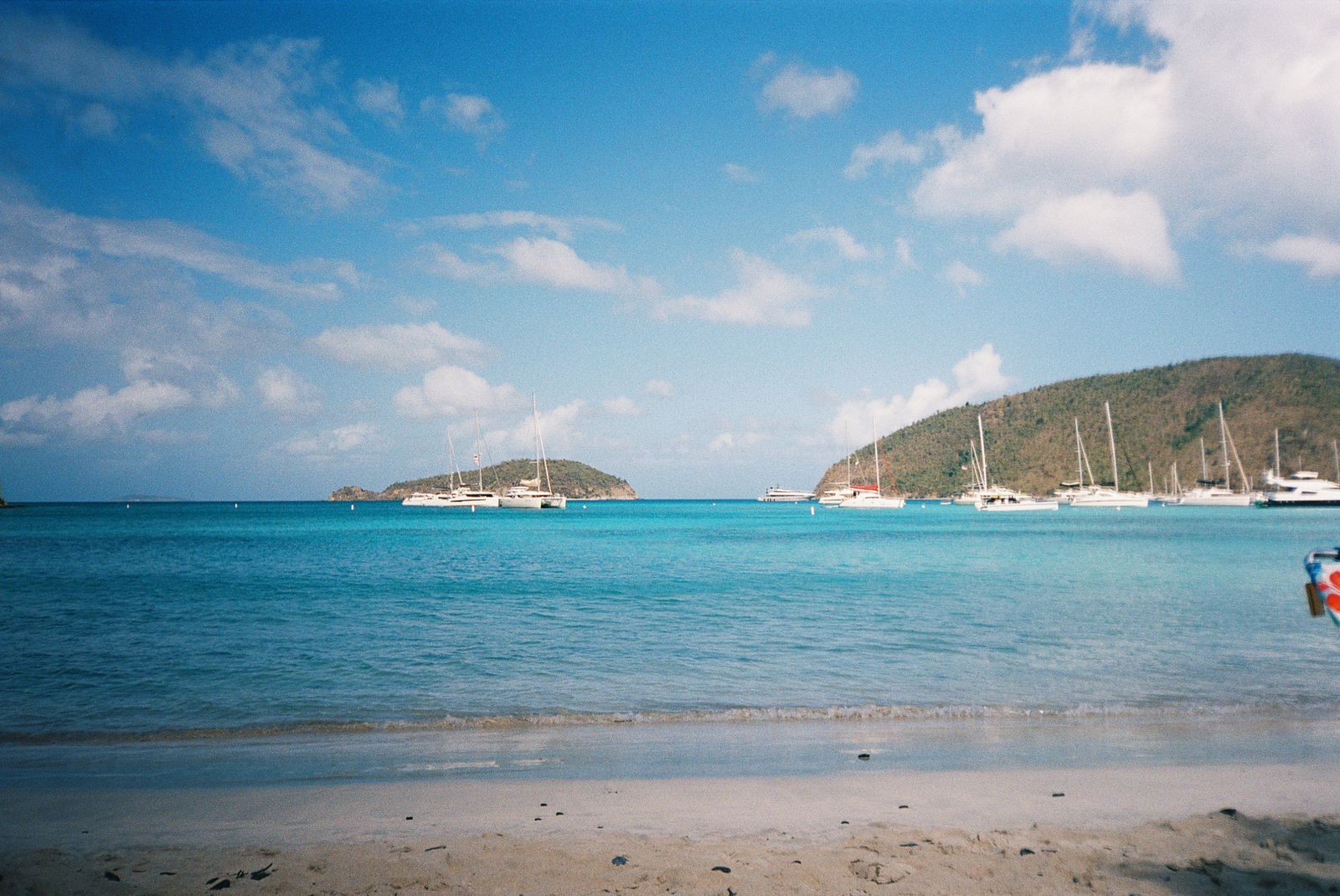Catamarans and sailboats anchored in turquoise water off a sandy beach, with a green hillside and a small island beyond